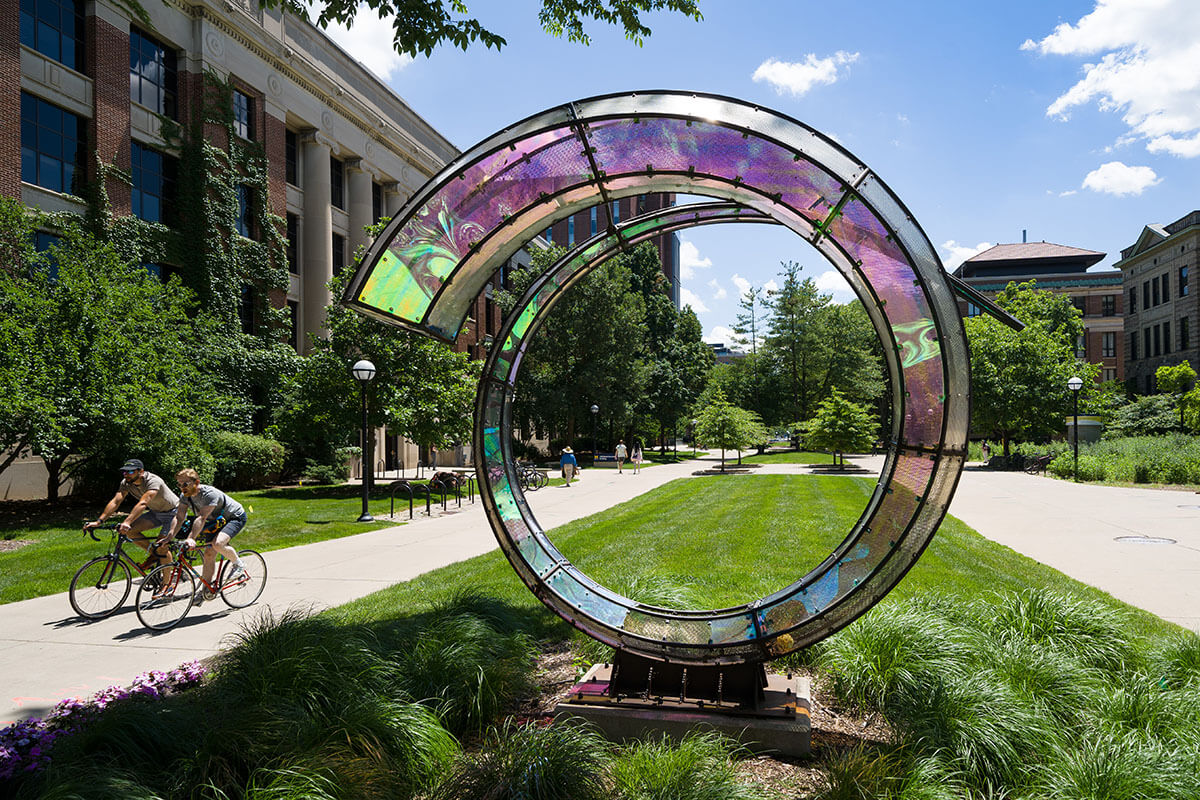 Students biking past the glass Arriving Home sculpture on Central Campus